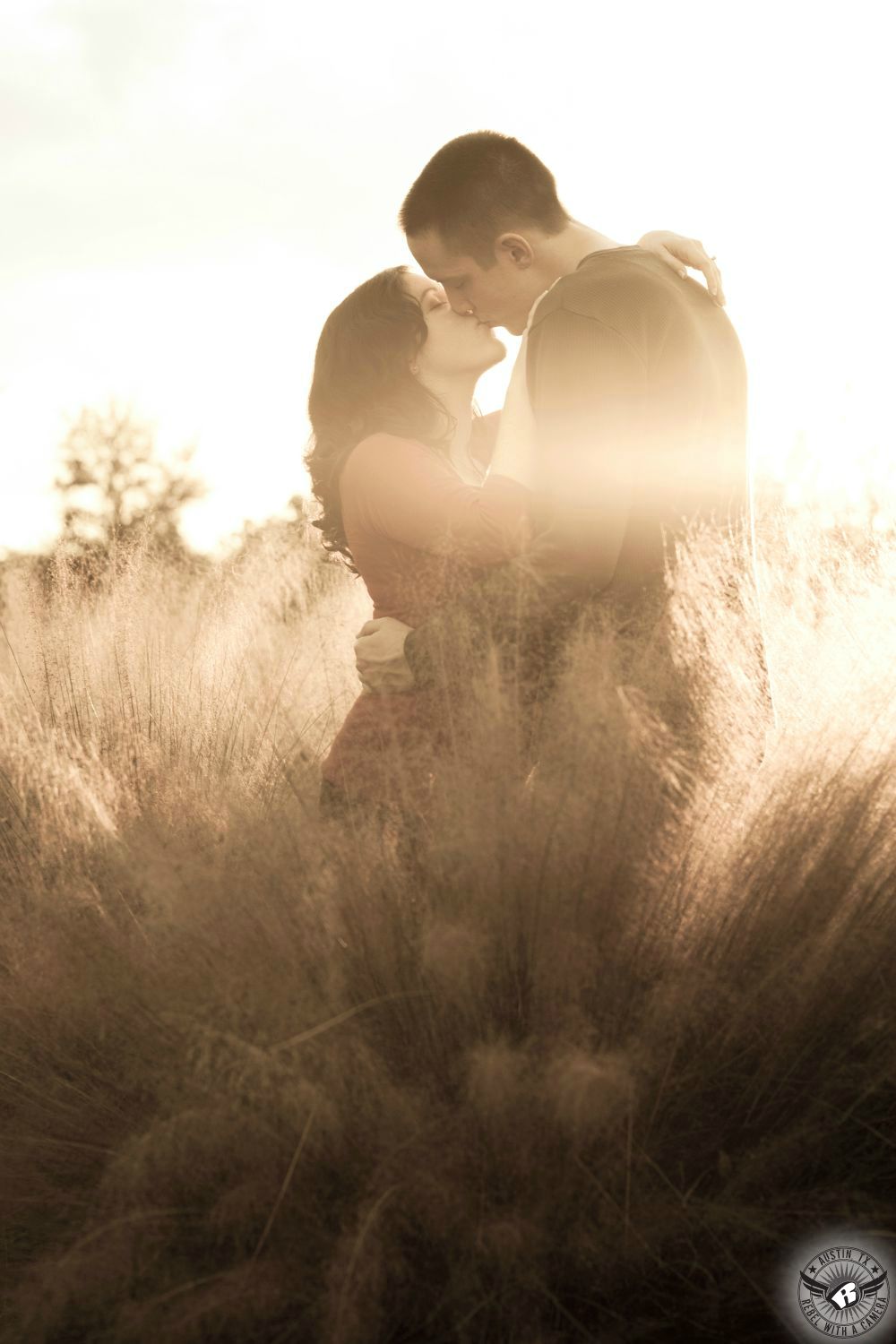 wavy haired brunette girl in a pink large weave sweater kisses a guy with dark short hair wearing a green long sleeve shirt while standing in fluffy sunlit brush with the sun warming the background in this Nicholas Sparks inspired engagement photograph in Austin.